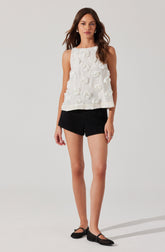 A woman is wearing a sleeveless white top with 3D floral embellishments and a round neckline. She pairs it with high-waisted black shorts that have a textured appearance. She is also wearing black slip-on shoes with a small heel and ankle straps.